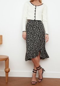 Cardigan blanc à manches longues avec des boutons noirs, associé à une jupe midi noire ornée de motifs floraux blancs et d'un ourlet à volants. Talons noirs à lanières.