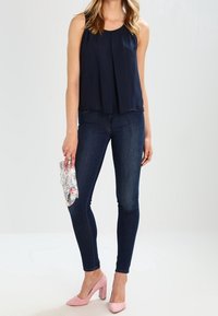 Marinblå ärmlös topp med veck, kombinerad med mörka skinny jeans och ljusrosa klackskor med blockhæl. Håller en blommönstrad scarf.