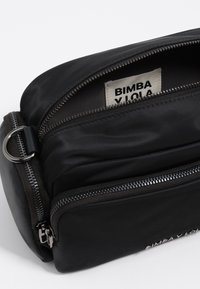 Čierna crossbody taška z textilu s uzatvárateľnými priehradkami, striebornými kovovými doplnkami a viditeľnou štítkom s logom vo vnútri. Hladká textura a kompaktný dizajn.