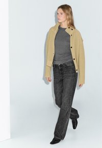 Cardigan beige a coste con bottoni neri, camicia grigia attillata e jeans a gamba larga grigio scuro. Stivali neri a punta completano l'outfit.