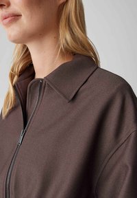 Braune Jacke mit Reißverschluss und markantem Kragen, gefertigt aus glattem Stoff mit leichtem Glanz, vorne mit einem Metallreißverschluss versehen.