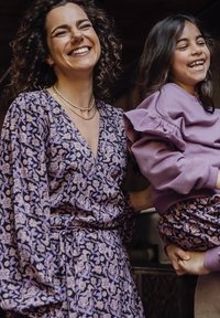 Bloemenpatroon jurk op een donkere achtergrond met paarse en crèmekleurige accenten; bijpassende paarse outfit met gerimpelde details gedragen door een kind.