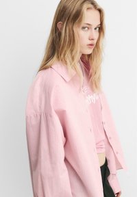 Camicia a righe rosa chiaro con bottone, dal taglio comodo e maniche oversize, indossata sopra una t-shirt grafica rosa. Tessuto di finitura liscia.