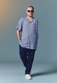 Chemise à manches courtes avec un motif bleu et blanc, pantalon marine et baskets blanches, accessoirisée avec des lunettes de soleil rondes.