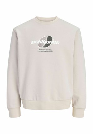 Garçon blond debout, portant une veste à capuche noire, un sweat-shirt blanc avec un motif graphique, un jean large foncé et des baskets noires sur un fond clair uni.