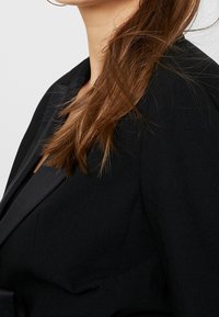 Blazer negro con un diseño estructurado, textura suave y solapas con muesca, que cuenta con un cierre de un solo botón y hombreras.