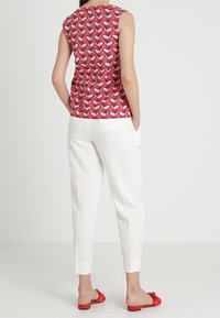 Ärmelloses rotes Oberteil mit geometrischem Muster, kombiniert mit weißen maßgeschneiderten Hosen und roten flachen Sandalen. Glatter Stoff, taillierte Passform.
