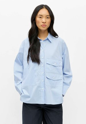 Jeune femme aux longs cheveux noirs portant une chemise à rayures bleu clair oversize avec de grandes poches frontales et un pantalon foncé.