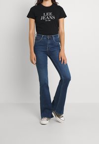 Camiseta de algodón negra con texto blanco "LEE JEANS EST. 1889", combinada con unos jeans acampanados de talle alto en azul y zapatillas de deporte con estampado.