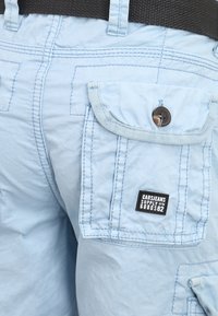 Pantaloni cargo azzurro chiaro realizzati in tessuto con una superficie liscia, dotati di numerose tasche cucite e una toppa nera con logo.