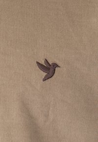 Beige corduroy stof med en lille, mørkebrun broderet kolibri på venstre side. Subtil ribbet tekstur synlig.