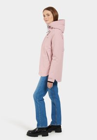 Ljusrosa vattentät jacka med huva, med justerbara snoddar, två framfickor och en avslappnad passform. Matchad med blå jeans och svarta stövlar.