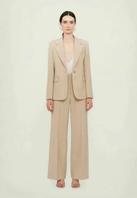 Femme se tenant debout, vêtue d'un blazer beige cintré et d'un pantalon large, avec un haut blanc transparent et des talons ouverts beige, sur un fond uni.