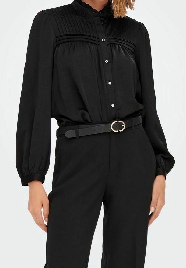 Femme portant un chemisier noir à manches longues avec des plis et des boutons, rentré dans un pantalon noir taille haute avec une ceinture noire et une boucle dorée.
