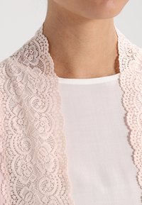 Spetschals i mjuk rosa med invecklade blommönster. Den genomskinliga tyget hänger lätt över en vit topp och framhäver texturer och detaljer.