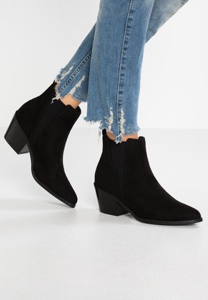 Benedenste benen in lichtblauwe gescheurde jeans gecombineerd met zwarte suède enkellaarsjes met puntige neuzen en blokhakken.