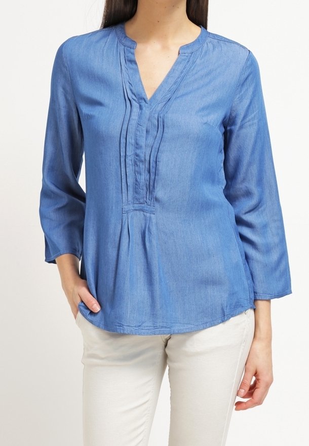 Niebieska bluzka z denimu z rękawami o długości trzech ćwiartek, dekoltem w kształcie litery V, plisowanym przodem i zaokrąglonym dołem, wyeksponowana na jasnych spodniach.