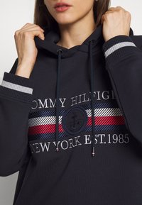 Marinblå hoodie av bomull med dragsko i huvan. Har en logotypapplikation och randiga detaljer i röd, vit och blå framtill.