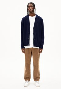 Cardigan en tricot navy avec six boutons, porté sur un t-shirt blanc, associé à un pantalon en velours côtelé marron et des baskets blanches. Design simple et texturé.