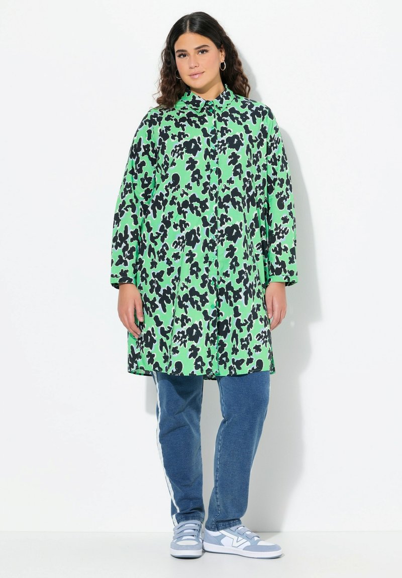 Cappotto lungo verde con motivo astratto nero, chiusura a bottoni e maniche lunghe. Abbinato a jeans blu e sneakers azzurri.