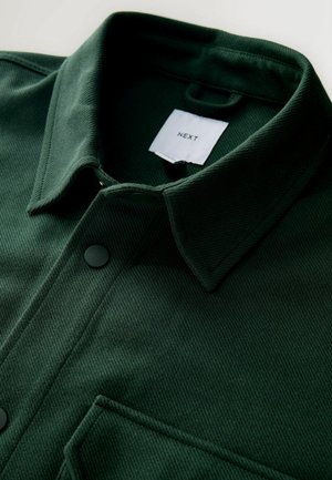 Cămașă verde închis cu nasturi, confecționată dintr-un material texturat, cu un design nervurat, guler ascuțit și buzunar la piept cu nasture negru.