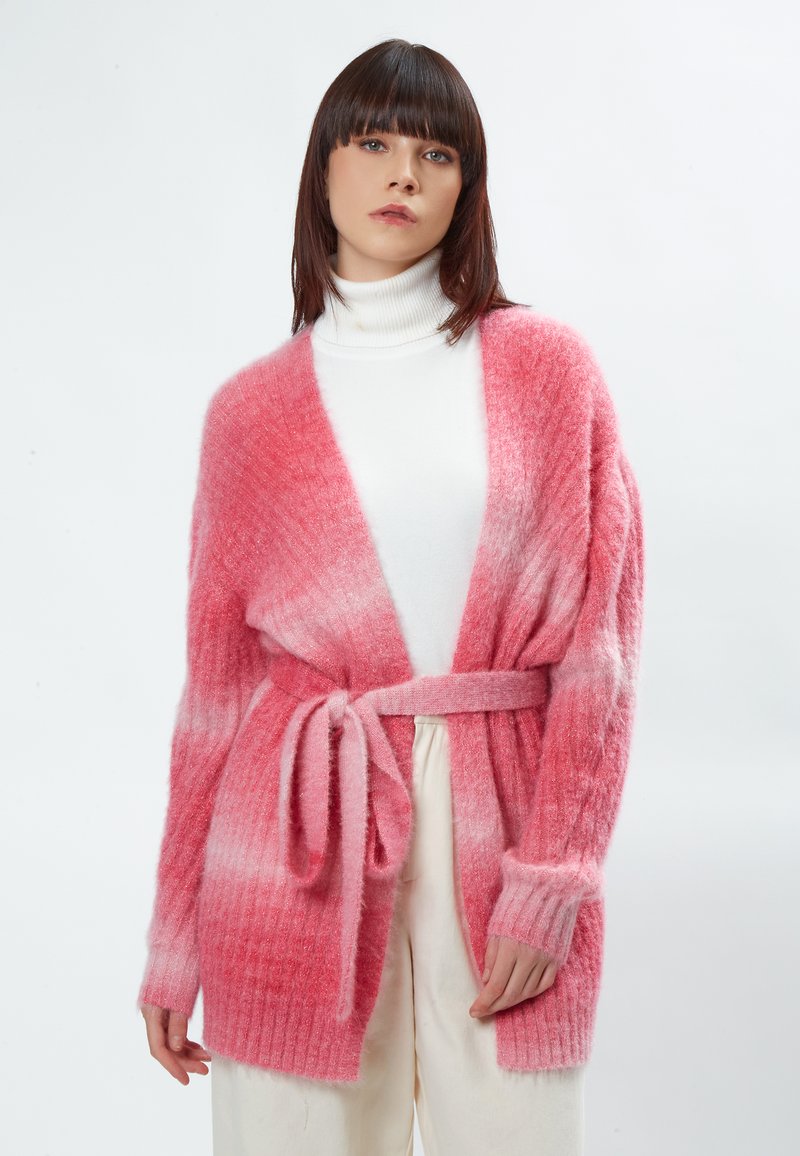 Roze gradient cardigan met geribbelde textuur en lange mouwen, voorzien van een strikceintuur in de taille, gedragen over een witte coltrui.