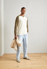 Pikkade varrukatega, kreemja ja oliivroheline pesapallisärk logoga, helesinised teksad ja rohelise ning valgega tennisetossud. Beez mantel hoitakse käes.