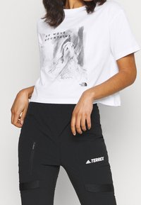 Camiseta blanca corta con un estampado gráfico gris y texto negro. Combinada con pantalones negros que cuentan con cremalleras laterales y un detalle de logo.