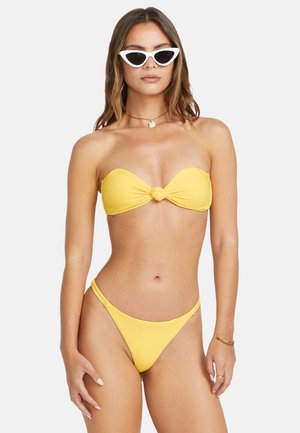 Bikini bandeau amarillo con detalle de nudo en el centro; textura acanalada en la tela; combinado con braguitas de bikini delgadas a juego. Gafas de sol blancas estilo cat-eye.