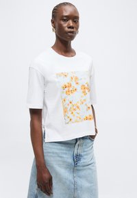 Γυναίκα που φοράει λευκό κοντομάνικο πουκάμισο με πορτοκαλί λουλουδάτο σχέδιο και ανοιχτό μπλε τζιν φούστα, στέκεται με το ένα χέρι στην τσέπη.