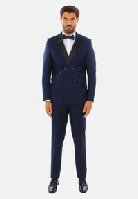 Tuxedo doppiopetto blu navy con rever in satin, pantaloni abbinati, camicia bianca e cravatta a farfalla. Scarpe eleganti nere lucide.