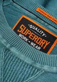 Pullover in cotone color teal con collo a coste. Presenta un'etichetta nera e arancione con la scritta "SUPERDRY WORK-WEAR" e "QUALITÀ."
