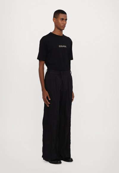 Camiseta negra de manga corta con un logo frontal, combinada con pantalones negros holgados y plisados, y calzado negro. Diseño y textura minimalistas.