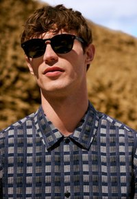 Giovane uomo con capelli castani che indossa occhiali da sole neri e una camicia a quadri blu con bottoni all'aperto con uno sfondo roccioso sfocato.