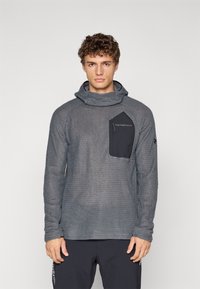 Grå teksturert langermet topp med cowl-hals og en svart brystlomme med logo. Stoffet har et vevd mønster og en avslappet passform.