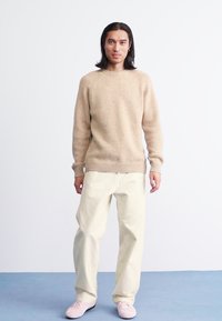 Beige gestrickter Pullover mit geripptem Saum, kombiniert mit cremefarbenen, lockeren Hosen und hellrosanen Sneakers, getragen von einer stehenden Person.