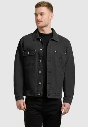 Mand iført sort denimjakke med knapper og brystlommer, stående mod en ensartet lys baggrund, kiggende til siden.