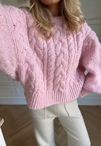 Pull en maille torsadée rose avec une texture épaisse, coupe oversize et ourlet côtelé, associé à un pantalon large couleur crème.