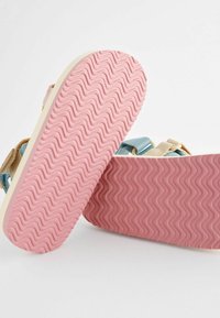 Sandal mit einer hellen Sohle in wellenförmiger rosa Textur, ergänzt durch blaue und beige Riemen. Einstellbare Verschlüsse sind enthalten.