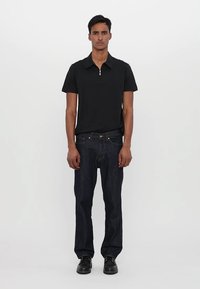 Polo preto de mangas curtas com gola com fecho, combinado com jeans de perna reta azul escuro e sapatos pretos, em pé sobre um fundo simples.