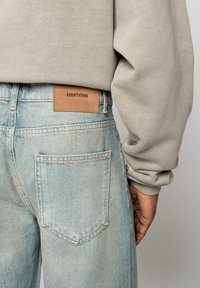 Personne portant un jean en denim bleu clair avec un patch en cuir marron étiqueté "EIGHTYFIVE" et un haut à manches longues beige, main visible sur le côté.