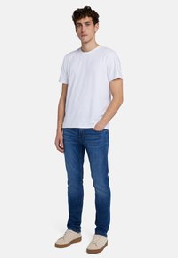 T-shirt bianca in cotone a maniche corte, abbinata a jeans di denim blu e sneakers beige, in piedi su uno sfondo bianco uniforme.