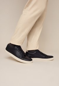 Schwarze Ledersneaker mit weißen Sohlen, getragen mit beigen Hosen und schwarzen Socken, Füße auf hellbeigem Hintergrund platziert.