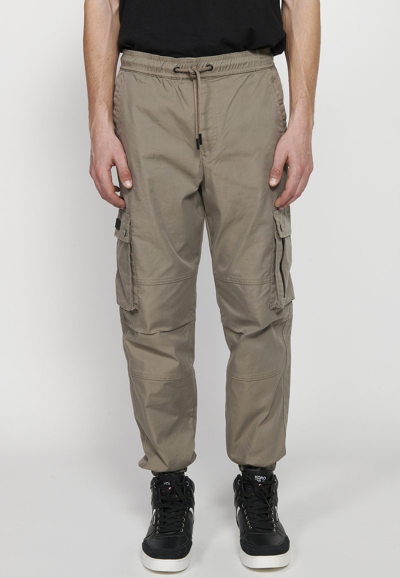 Beige cargo nadrág rugalmas derékpánttal, zsinóros részletekkel, több oldalsó zsebbel, szűkített lábformával és könnyű anyagból készült.