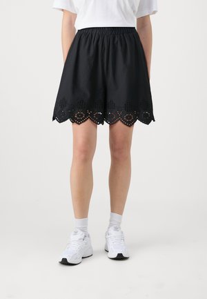 Zwarte rok met een elastische tailleband, gekartelde zoom en laser-geperforeerd oogjesdesign. Gestyled met een witte t-shirt en witte sneakers.