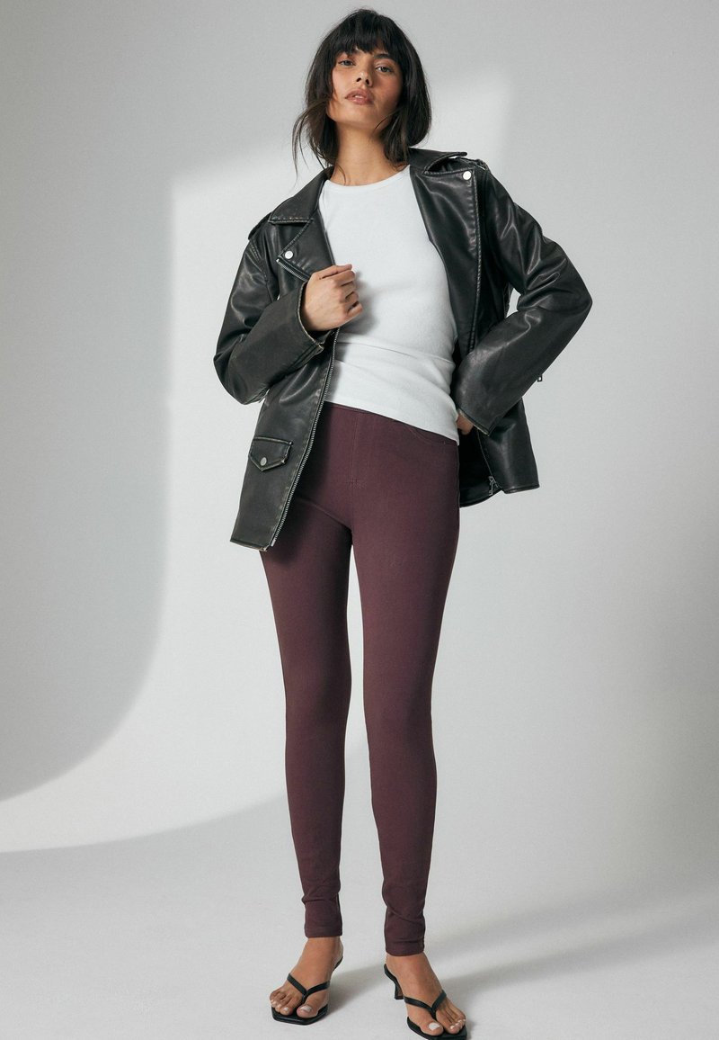 Chaqueta de cuero negra, camiseta blanca de manga larga y leggins burdeos con una textura suave; el modelo lleva sandalias negras de tiras.