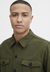 Olive green button-up shirt with a collar, two chest pockets, and black buttons. Made from sturdy fabric with a smooth texture.