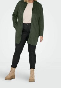 Abrigo largo verde de ante con apertura frontal, sobre una blusa beige, combinado con pantalones pitillo negros y botines beige con cordones y suelas gruesas.