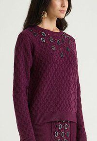 Pull en maille violet avec motif texturé, présentant des boutons décoratifs noirs et rouges autour de l'encolure. Poignets et ourlet côtelés.