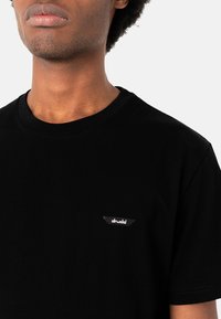 Black cotton T-shirt with a crew neck, featuring a small white logo patch on the left chest area and short sleeves. Simple design.